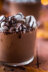 A close-up of a glass of hot chocolate topped with marshmallows and a drizzle of chocolate sauce, with autumn leaves in the background.
